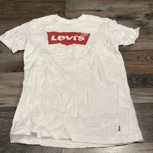 Vit Levi's t-shirt med röd logga - Vit t-shirt från Levi's med klassisk röd logga tryckt på bröstet. T-shirten är i bomull, har rund hals och korta ärmar. Enkel och stilren modell som passar perfekt till jeans eller shorts.