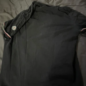 Svart Moncler t-shirt - Svart t-shirt från Moncler med klassisk logga på ärmen och snygga ränder i vitt och rött vid ärmslutet. Enkel och stilren design som passar perfekt till jeans eller shorts. Gjord i mjuk bomull för skön känsla hela dagen.