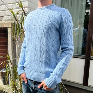 Ljusblå kabelstickad tröja Polo Ralph Lauren - Kabelstickad tröja från Polo Ralph Lauren i en snygg ljusblå färg. Klassisk rund halsringning, ribbade muddar och nederkant. Liten broderad logga på bröstet. Perfekt för dig som gillar en stilren och tidlös look.