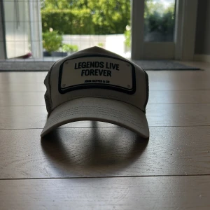 Beige truckerkeps John Hatter & Co - Snygg beige truckerkeps från John Hatter & Co med svart mesh på sidorna och bak. Framtill sitter en vit patch med texten 'Legends Live Forever' i svart. Böjd skärm och justerbar passform. Perfekt för dig som vill sticka ut med en cool accessoar.
