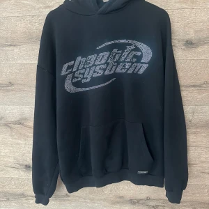 Svart hoodie Cheap Monday med tryck - Svart hoodie från Cheap Monday med stort silvrigt tryck på bröstet där det står 'cheap tile system'. Klassisk känguruficka framtill, huva och ribbade muddar. Perfekt för dig som gillar streetwear och vill ha en chill look.