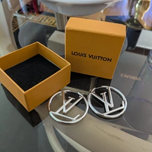 Louis Vuitton silverörhängen LV-logo - Stilrena örhängen i silverfärgad metall från Louis Vuitton. De är runda hoops med den ikoniska LV-loggan i mitten, perfekta för att lyfta vilken outfit som helst. Kommer i originalask.