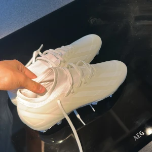 Vita fotbollsskor från Adidas - Snygga helvita fotbollsskor från Adidas med klassiska tre ränder på sidan. Skorna har snörning, rund tå och platt sula med dobbar för gräsplan. Ovandelen är i syntetmaterial och innersulan är mjuk för extra komfort. Perfekta för dig som vill sticka ut på planen. ALLDRIG använt pga storlek