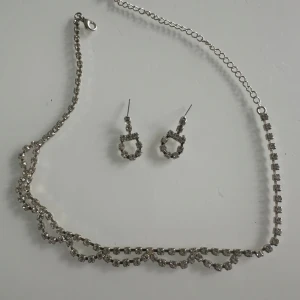 Glittrigt halsband och örhängen set - Snyggt set med halsband och matchande örhängen i silverfärg med glittrande stenar. Halsbandet har dubbla rader av stenar som ger extra bling och örhängena är små hoops med samma gnistrande look