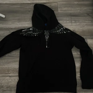 Marcelo Burlon svart hoodie med vingar - Svart hoodie från Marcelo Burlon County of Milan med ikoniskt vingtryck i vitt och grått över axlar och bröst. Tröjan har huva med snörning och långa ärmar. Perfekt statement-plagg för dig som vill sticka ut.