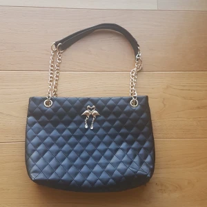 Svart quiltad axelväska med kedja - Snygg svart axelväska i quiltat skinnimitation med guldfärgade kedjor och ett dekorativt emblem med två svanar och gnistrande detaljer framtill. Väskan har dragkedja upptill. Perfekt för dig som vill ha en stilren men ändå unik väska. PRIS KAN DISKUTERAS!