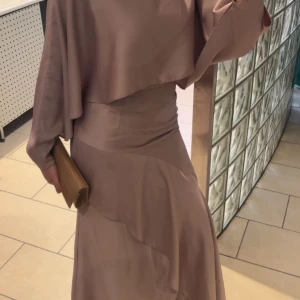 Ljuslila långklänning med vida ärmar - Säljer en elegant ljuslila långklänning i ett glansigt, mjukt material. Klänningen har vida, flödande ärmar och en cape-liknande överdel som ger en cool och unik siluett. Perfekt för dig som vill sticka ut med en stilren och modern look.