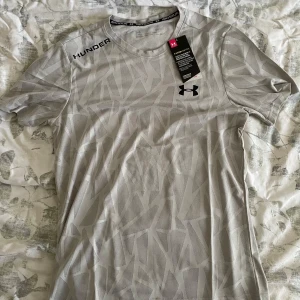 Grå mönstrad t-shirt från Under Armour - Snygg grå t-shirt från Under Armour med diskret geometriskt mönster och svarta loggor på bröstet och axeln. Tillverkad i ett lätt och ventilerande syntetmaterial, perfekt för träning. Klassisk passform med korta ärmar och rund hals.