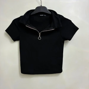 Svart ribbad polotopp med dragkedja - Svart ribbad polotopp från Busem med korta ärmar och croppad passform. Toppen har en cool silverfärgad dragkedja framtill med rund detalj i zippen. Storlek S men sitter som Storlek XS/XXS.