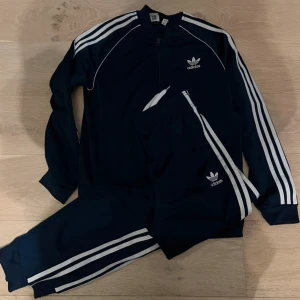 Adidas mörkblå träningsset storlek 176/small - Mörkblå träningsbyxor från Adidas med klassiska vita ränder längs sidorna och broderad logga på benet. Tillverkade i polyester och har en sportig, loose passform. Perfekta för dig som gillar streetwear och sportig stil.