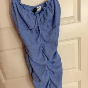 Blå kort omlottklänning med snörning - Supersöt blå kort klänning med smockat tyg, snörning vid bysten och snygg rynkdetalj framtill. Figurnära passform och ärmlös design, perfekt för varma dagar eller när du vill känna dig extra fin.