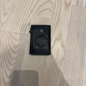 Svart kolfiberinspirerad plånbok (helt ny) - Slimmad svart plånbok med kolfiberlook och smart knappstängning. Plånboken har plats för kort och sedlar, och den stilrena designen gör den perfekt för dig som gillar minimalistiska accessoarer. Snygg detalj med struktur på ytan.