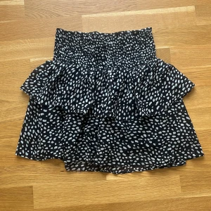 Svartvit volangkjol från Chelsea - Supersöt kort kjol från Chelsea i svart med vita prickar. Kjolen har bred smock i midjan och dubbla volanger som ger en riktigt flowy känsla. Perfekt för dig som gillar att sticka ut lite extra med ett lekfullt mönster. Finns en liten defekt längs ner på kjolen (bild 2) men inget man tänker på när man har på sig den tycker jag! Går alltid att sy in den om det är något du stör dig på! Annars väldigt bra skick.