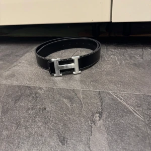 Svart skärp med H-spänne - Säljer ett svart skärp med ikoniskt H-spänne i silverfärgad metall. Bältet är gjort i präglat skinn med vita sömmar längs kanterna. Perfekt accessoar för att lyfta din outfit med en lyxig touch.