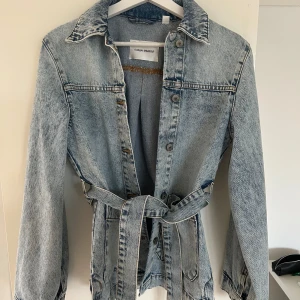 Ljusblå jeansjacka med bälte Carin Wester - Snygg ljusblå jeansjacka från Carin Wester med bälte i midjan och stora fickor framtill. Jackan har klassisk krage, knäppning med knappar och raka ärmar. Perfekt för dig som vill ha en chill och trendig look.