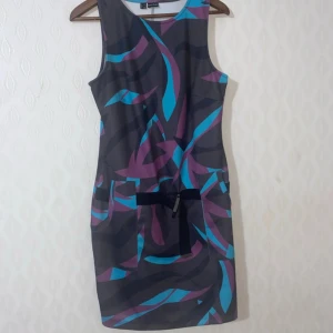 Super snygg helt ny klänning utan prislapp  - Snygg ärmlös kortklänning med abstrakt mönster i lila, blått och svart. Klänningen har två fickor framtill och ett svart knytband i midjan. Perfekt för dig som gillar färg och vill sticka ut lite extra.