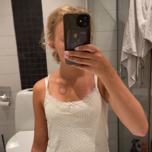 Vit spetskantad linnetopp Brandy Melville - Supersöt croppad vit linnetopp från Brandy Melville med små blå blommor, spetskant och en liten rosett framtill. Tunt och mjukt bomullsmaterial, smala axelband och figurnära passform. Perfekt för en chill och söt look.