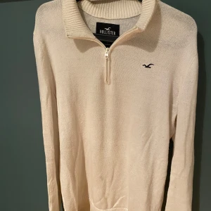 Beige stickad tröja från Hollister - Säljer en beige stickad tröja från Hollister med halv dragkedja och ribbad krage. Tröjan har lång ärm och diskret Hollister-logga på bröstet. Perfekt för lager-på-lager och chill stil.