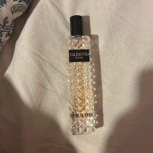 Valentino Donna Born In Roma parfym - Exklusiv parfym från Valentino, Donna Born In Roma. Flaskan är av genomskinligt glas med nitar och har en ljusrosa vätska inuti. Snygg svart etikett och svart kork ger en lyxig känsla. Perfekt för dig som gillar eleganta och trendiga dofter.