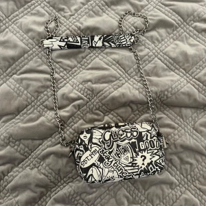 Guess axelväska med grafiskt tryck - Cool axelväska från Guess med svartvitt grafiskt mönster och text, kedjerem och detaljer i silver. Väskan har rektangulär form och dragkedja upptill. Perfekt för dig som gillar streetstyle och vill sticka ut med en unik accessoar.