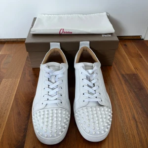 Vita Christian Louboutin sneakers med nitar - Snygga vita sneakers från Christian Louboutin med ikoniska röda sulor och coola vita nitar på tån. Skorna har snörning och är tillverkade i skinn för en lyxig känsla. Perfekta för dig som vill sticka ut med en exklusiv och edgy stil. Använda en gång och pris kan diskuteras
