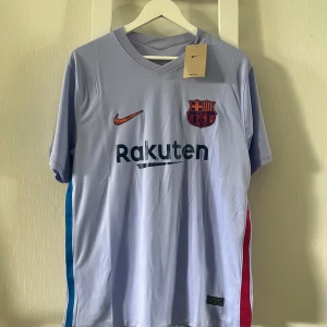 FC Barcelona bortatröja Nike M - FC Barcelona bortatröja från Nike i ljusblått med klubbmärke och orange Nike-logga på bröstet. Rak passform, kortärmad och tillverkad i lätt, ventilerande polyester. Sidorna har blå och röda detaljer. Rakuten och UNICEF-loggor tryckta på fram- och baksidan.