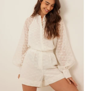 Vitt set med shorts och blus - NA-KD - Snyggt set med högmidjade shorts från NA-KD i ett lätt, halvtransparent tyg med subtilt strukturmönster. Shortsen har breda ben, uppvikta benslut och dold dragkedja i sidan.  Blusen har vita knappar och ballongärm. Storlek 38 i båda. Går att köpa separat om man önskar det🧡