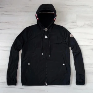 Svart vindjacka från Moncler med huva - Svart vindjacka från Moncler med huva och dragsko, hel dragkedja och två fickor framtill. Jackan har Moncler-logga på ärmen och snygga röd-vita detaljer på huvan. Perfekt för dig som vill ha en stilren look.