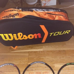 Wilson Tour tennisväska svart/orange - Stor Wilson Tour tennisväska i svart med orange detaljer och logga i gul, orange och röd. Väskan har flera fack med dragkedja och breda, vadderade axelremmar i orange. Perfekt för att bära flera racketar och tillbehör till tennisbanan.