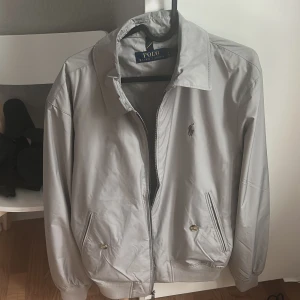 Grå vindjacka från Polo Ralph Lauren - Snygg grå vindjacka från Polo Ralph Lauren med klassisk krage, dragkedja framtill och två fickor med knapp. Jackan har en stilren look med diskret logga på bröstet och ribbade muddar vid ärmslut och midja. Perfekt för dig som gillar enkel och clean stil . Kan diskutera om priset på dm