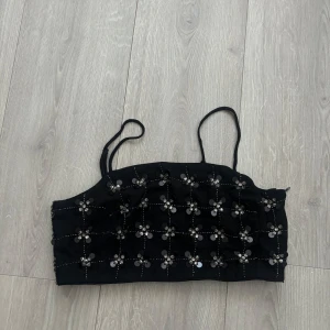 Svart paljett-topp från & Other Stories - Supersnygg svart topp från & Other Stories Paris Atelier med smala axelband. Toppens framsida är täckt av blommiga paljettdetaljer och små stenar som ger en glittrig effekt. Perfekt för att sticka ut och ge outfiten extra edge.