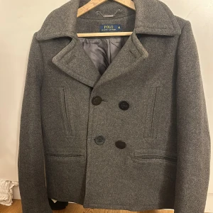 Grå kappa från Polo Ralph Lauren - Klassisk grå skepparkavaj från Polo Ralph Lauren med dubbelknäppning och bred krage. Jackan är i ullblandning och har raka fickor framtill samt helfodrad insida. Perfekt för dig som gillar en tidlös och stilren look under höst och vinter. Små hål inuti vid armarna men syns ej när man har på sig. Orginalpris:4000 säljer för 950kr