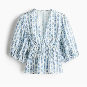 Blommig blus med puffärm - Supersöt blus i vitt med blått blommönster, djup v-ringning och smockad midja. Puffiga trekvartsärmar ger en romantisk vibe. Perfekt för dig som gillar en feminin och trendig look.