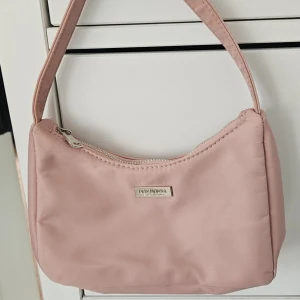 Rosa axelväska från Don Donna - Söt ljusrosa axelväska från Don Donna med stilren design. Väskan har ett huvudfack med dragkedja och en smidig innerficka. Tillverkad i slät polyester och har ett diskret metallogo framtill. Perfekt storlek för det viktigaste du behöver ha med dig.
