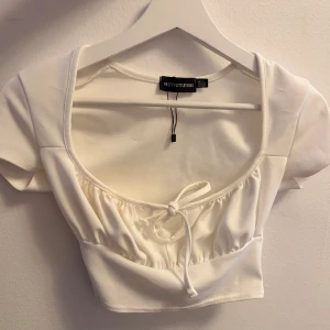 Vit croppad topp PrettyLittleThing - Superfin vit croppad topp från PrettyLittleThing med korta ärmar och söt knytning framtill. Toppen har en fyrkantig ringning och lätt rynkade detaljer vid bysten. 