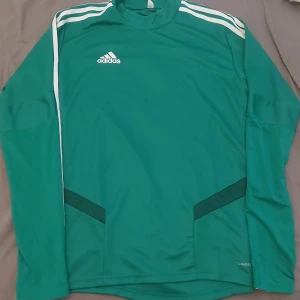Grön långärmad Adidas träningsoverall storlek Small. climacool - Adidas Tiro 19 långärmad fotbollströja i grön med vita ränder på axlarna och ärmarna. Tröjan har klassisk Adidas-logga på bröstet och är tillverkad i lätt, ventilerande ClimaCool-material. Perfekt för träning eller match.
