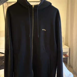 Svart hoodie med dragkedja från BOSS - Snygg svart hoodie från BOSS med dragkedja framtill och diskret logga på bröstet. Tröjan har huva med snörning och långa ärmar. Perfekt för en chill och stilren look. Materialet känns mjukt och bekvämt, och passformen är normal.