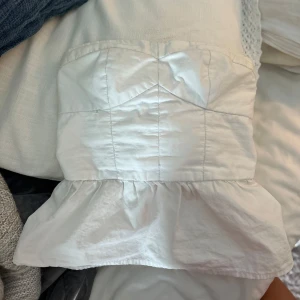Vit bandeau tubtopp med volang - Supersöt vit bandeau tubtopp med volangkant nertill. Toppen har smockad rygg för extra stretch och sitter snyggt på kroppen. Perfekt för varma dagar och enkel att styla med jeans eller kjol.