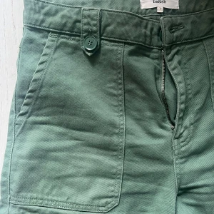 Gröna raka byxor från ba&sh - Snygga gröna byxor från ba&sh med raka ben och hög midja. Byxorna har stora fickor fram och bak, stängs med knapp och dragkedja. Tillverkade i mjuk bomull, perfekta för en chill och stilren look.