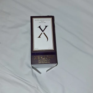 Xerjoff Accento Eau de Parfum 100ml - Lyxig parfym från Xerjoff, Accento, i en unik lila sammetsflaska med gulddetaljer och elegant guldlock. Flaskan rymmer 100 ml och kommer i en snygg mörklila kartong med guldfärgade inslag. Perfekt för dig som gillar exklusiva dofter och snygg design.