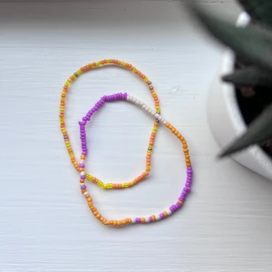 Färgglada pärlarmband i lila och orange - Två tunna armband med små pärlor i lila, orange, vitt och gult. Somrig känsla med mixade färger i olika nyanser. Perfekta för att addera lite färg till din stil. Armbanden är elastiska, gjorda av gummiband. Önskar du veta exakta mått? Tveka inte att höra av dig! 💜🧡🤍