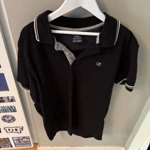 Svart pikétröja från Champion - Svart pikétröja från Champion med grå detaljer på insidan av kragen och vita ränder på krage och ärmslut. Klassisk passform och liten Champion-logga på bröstet. Perfekt för en clean och sportig stil.