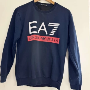 Mörkblå EA7 Emporio Armani sweatshirt - Snygg mörkblå sweatshirt från EA7 Emporio Armani med stor vit och röd logga framtill och tryck på ärmen. Tröjan har rund hals, ribbade muddar och är tillverkad i mjuk bomull. Perfekt för dig som gillar stilrena plagg.