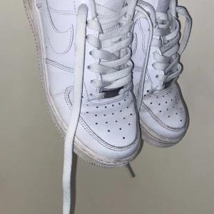 Nike Air Force 1 vita sneakers - Klassiska Nike Air Force 1 sneakers i helvitt skinn med perforerad tå och vit sula. Skorna har rund tå, snörning och den ikoniska swoosh-loggan på sidorna. Perfekta för en clean och stilren look.