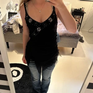 Svart linnetopp med spets och blommor - Svart linnetopp med v-ringning och smala axelband dekorerade med spets. Framsidan har blommiga detaljer och små pärlor som ger en cool look. Toppen har asymmetrisk nederkant i tunt, skirt material som ger extra edge.