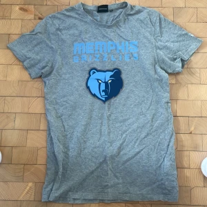 Grå Memphis Grizzlies t-shirt New Era - Snygg grå t-shirt från New Era med Memphis Grizzlies-tryck och björnmotiv i blått framtill. Klassisk passform och korta ärmar, perfekt för dig som gillar basket och streetwear. Tillverkad i mjuk bomull för skön känsla hela dagen.