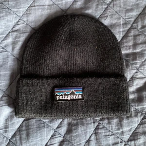 Svart ribbstickad mössa från Patagonia - Svart ribbstickad mössa från Patagonia med klassisk uppvikt kant och broderad logotyp framtill. Perfekt för kalla dagar och enkel att matcha med alla outfits. Mjuk och skön passform som sitter snyggt på huvudet.
