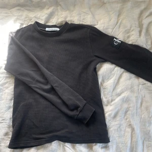 Svart långärmad tröja Calvin Klein Jeans - Svart långärmad tröja från Calvin Klein Jeans med diskret struktur i tyget och rund hals. På vänster ärm sitter en svart patch med CK-loggan. Tröjan har en klassisk passform och är perfekt för en clean och stilren look. Storlek S i bra skick!