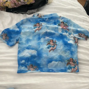 Blå t-shirt med änglamotiv - Säljer en unik blå t-shirt med moln och änglar tryckta över hela plagget. Skjortan har korta ärmar. Storlek står inte på plagget men uppskattningsvis XS-M
