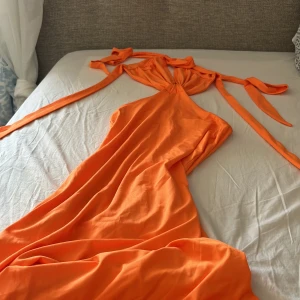 Lång orange klänning med knytband - Superfin långklänning i stark orange färg med snygga knytband vid axlarna och djup v-ringning. Klänningen har en flowy passform och är tillverkad i ett mjukt, lätt tyg som känns skönt mot huden. Perfekt för dig som vill sticka ut med färg och coola detaljer.